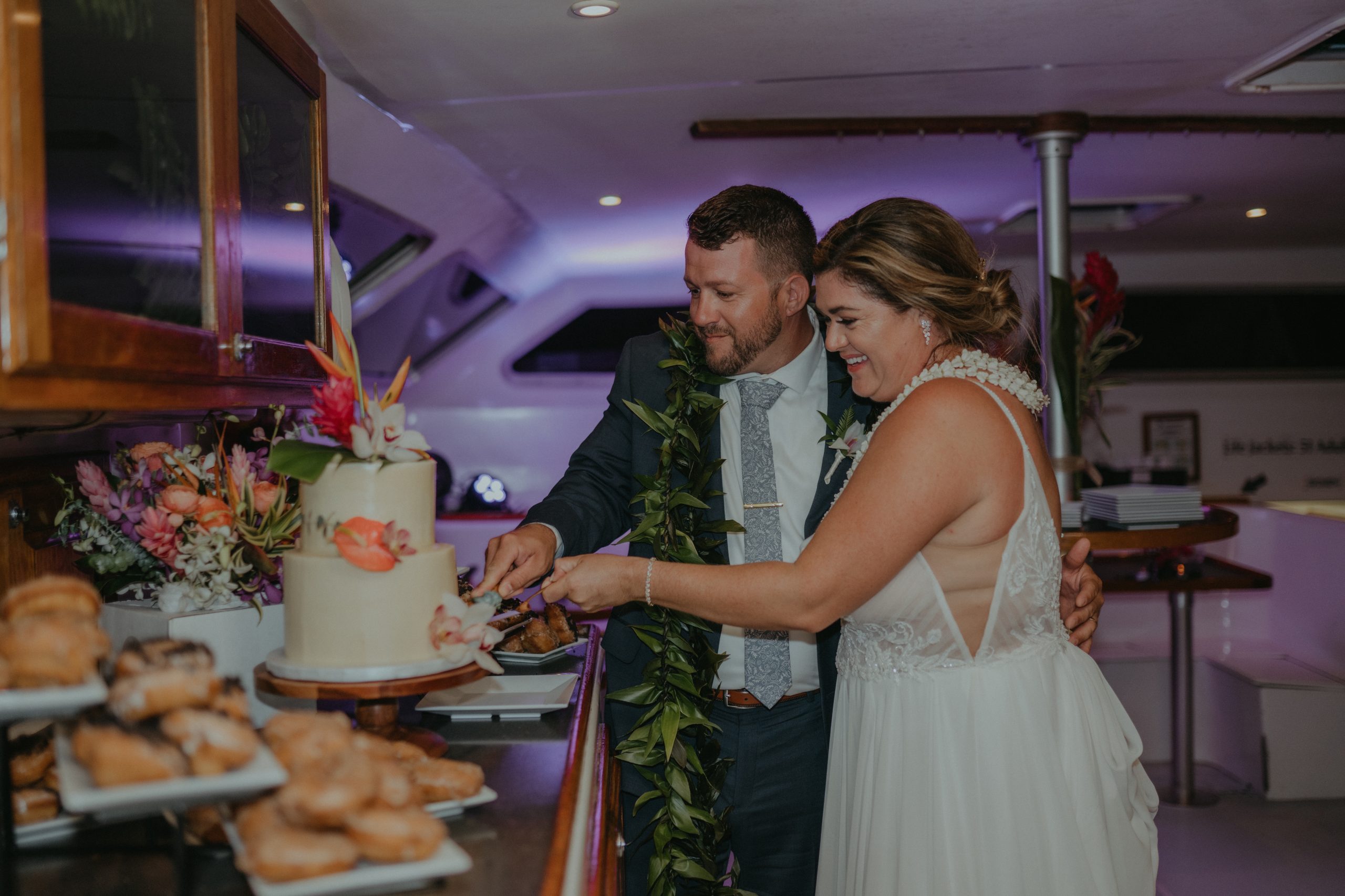Bride and groom cutting wedding cake on a yacht, surrounded by pastries and flowers.