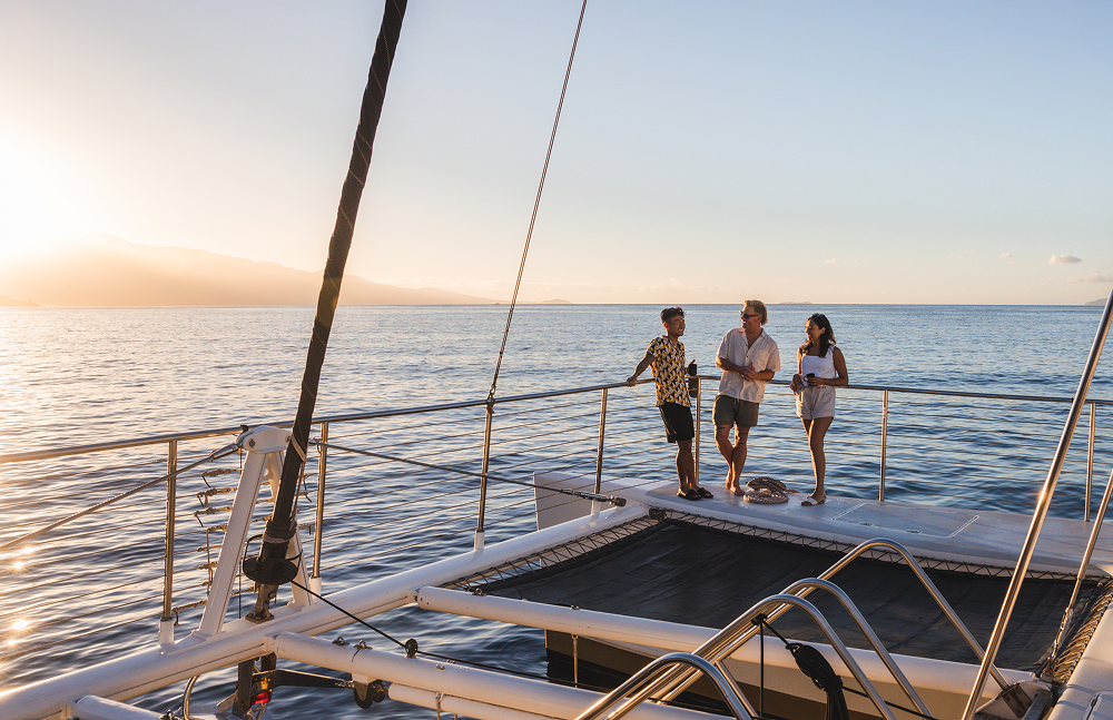 Three people standing on a catamaran enjoying a sunset over the ocean.