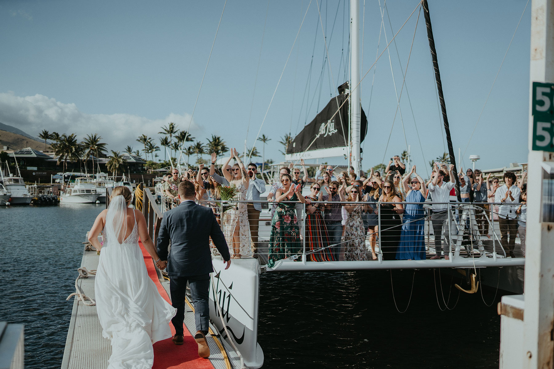 Bride and groom walking towards cheering crowd on a dock beside a sailboat.