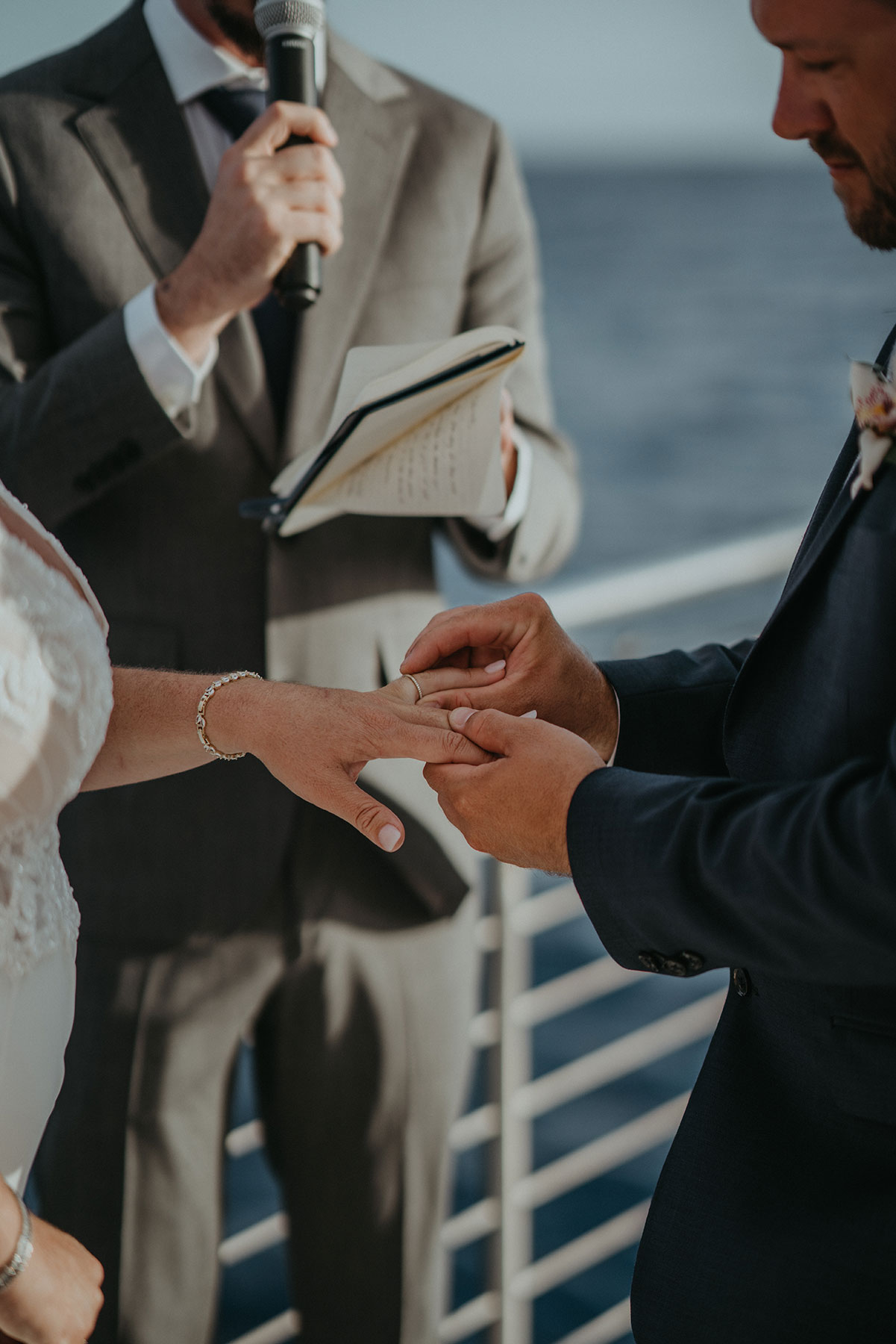 Couple exchanging rings during a wedding ceremony on a boat with officiant holding a microphone and book.