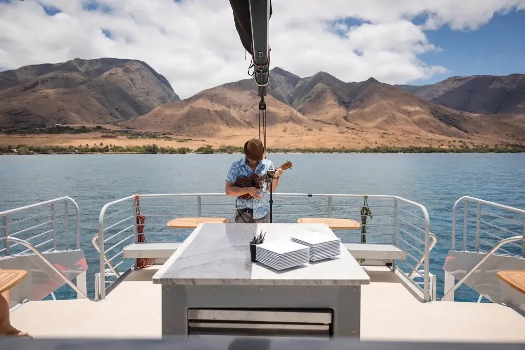 Person playing ukulele on a boat with mountain view in the background.