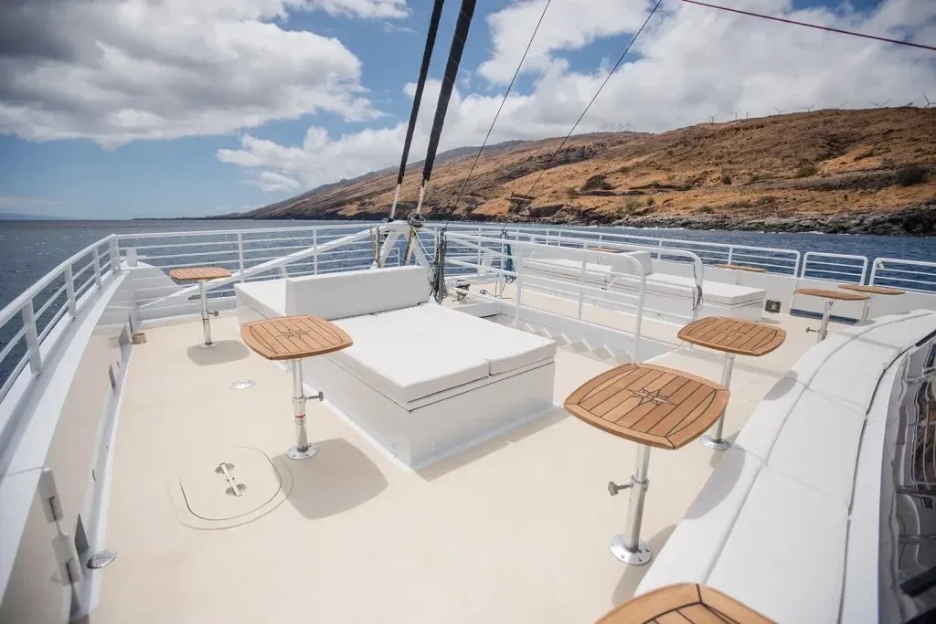 Boat deck with wooden stools, surrounded by railings, near a rocky coastline under a partly cloudy sky.