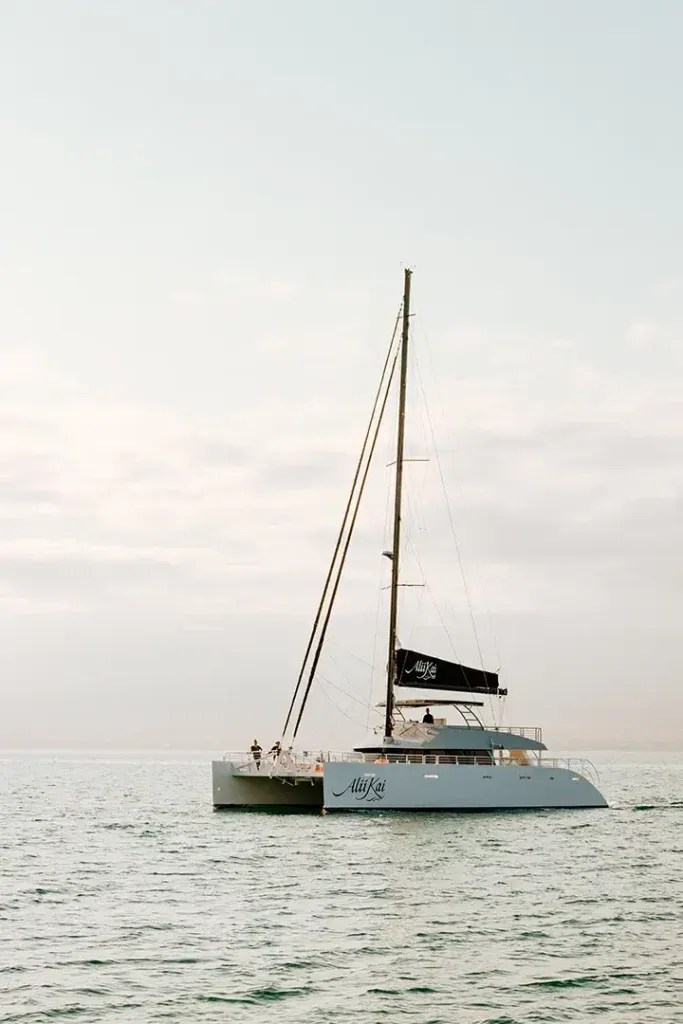 Sailboat on calm sea with cloudy sky, black sail, and several people aboard.