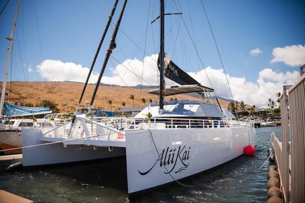 A white catamaran named Alii Kai docked in a marina on a sunny day.