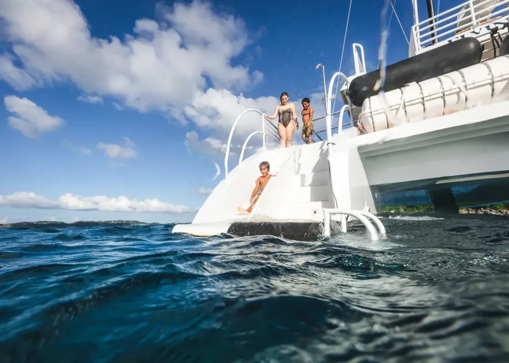 Children diving off a boat into the ocean under a blue sky with clouds.