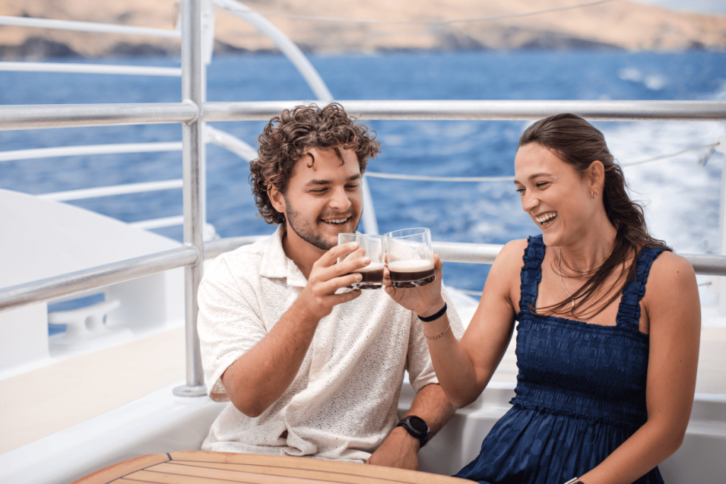 A man and woman clink glasses on a boat with ocean in the background.