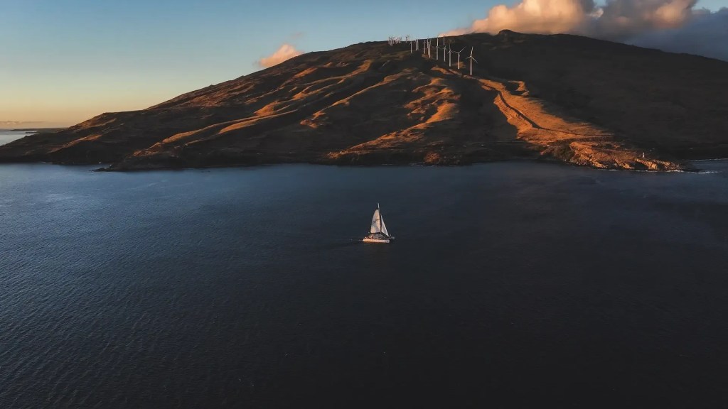 Sailboat on calm sea with hill and wind turbines in background at sunset.