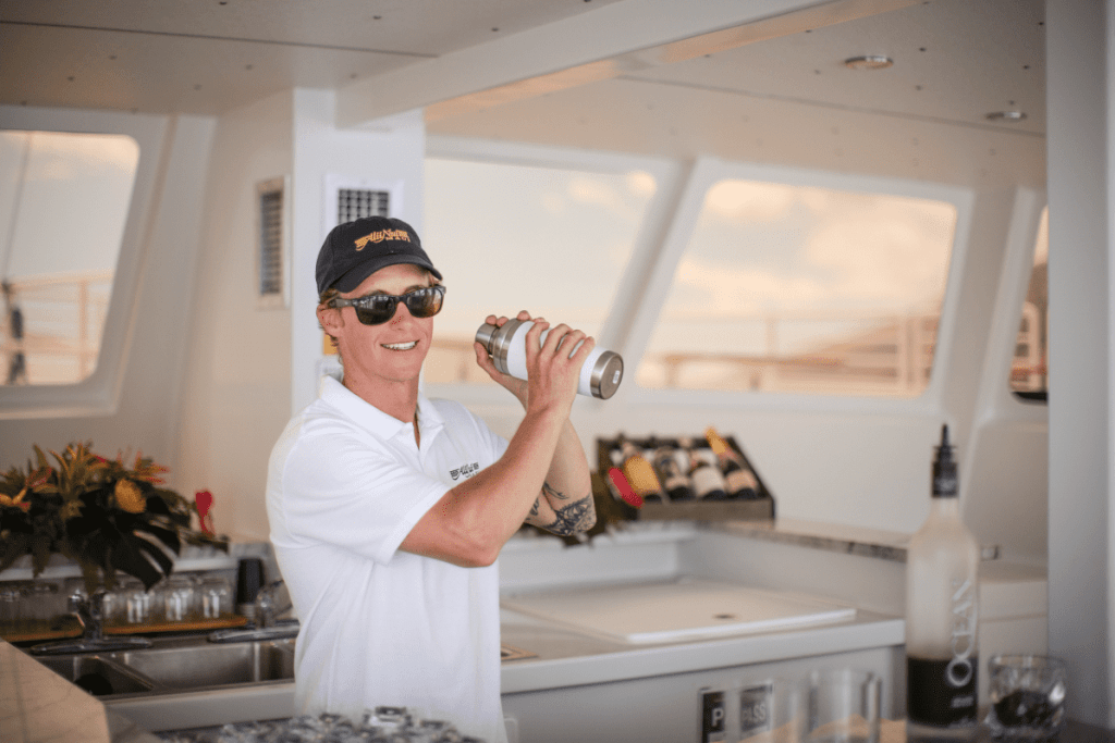 Person in sunglasses and cap shakes cocktail shaker on boat.