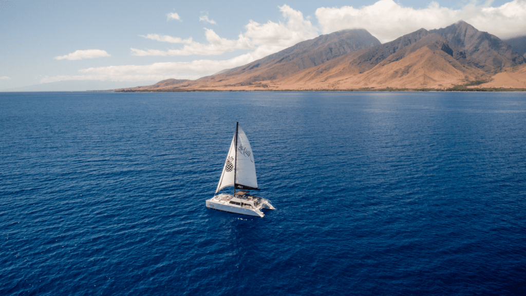 Sailboat on blue ocean near a mountainous coast under a partly cloudy sky.