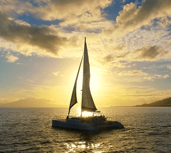 Sailboat on ocean at sunset with glowing sky and distant mountains.