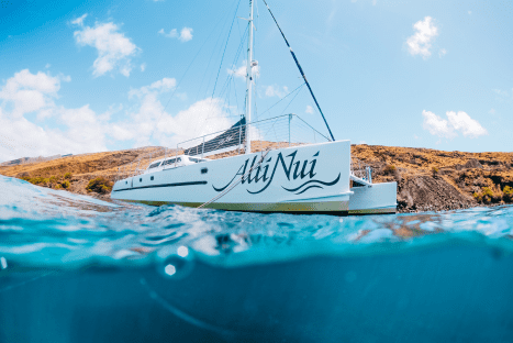 Catamaran named Alii Nui floating on the ocean near a rocky shore under a blue sky with clouds.
