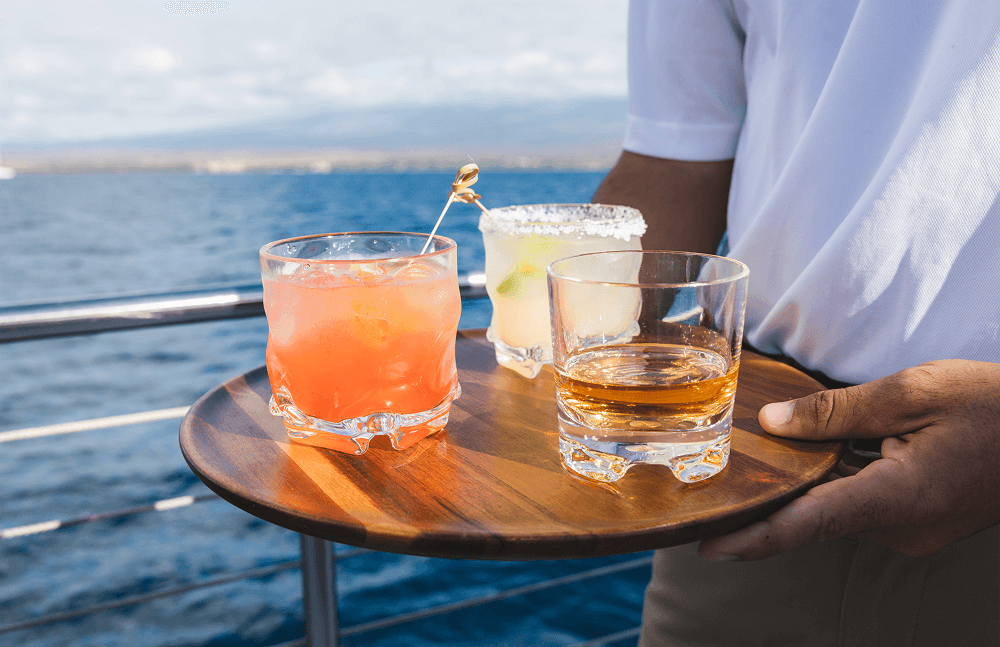 Person holding tray with three drinks on a boat with ocean view.