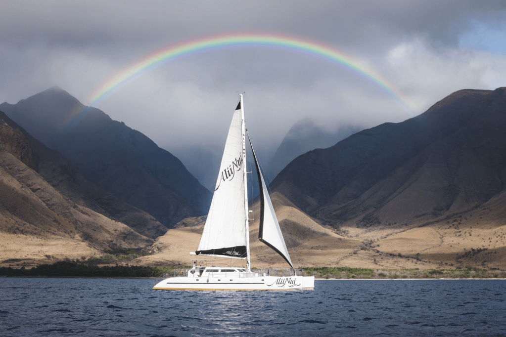 Sailboat on ocean with rainbow above mountains in the background.