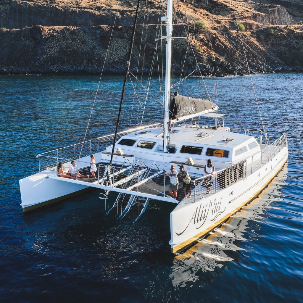 Large white catamaran sailing near a rocky coast with people on deck.