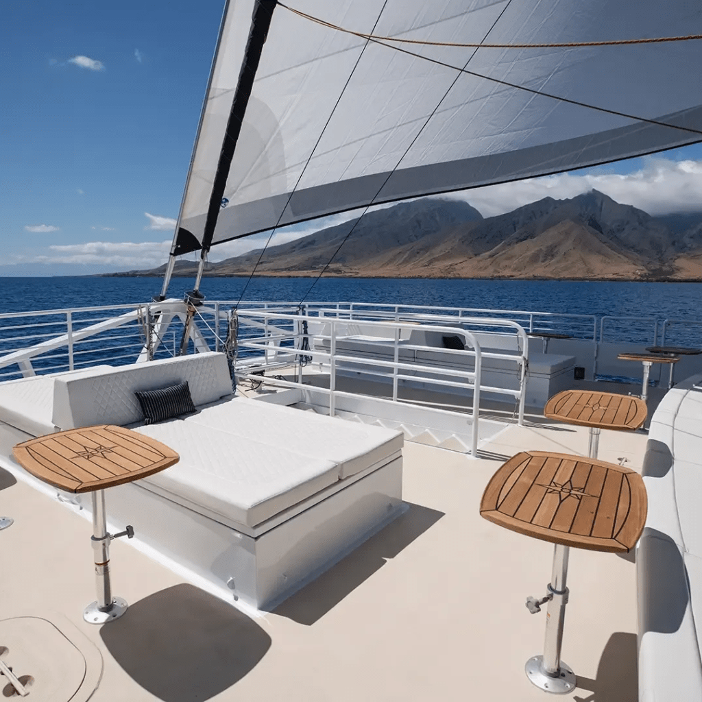 Sailboat deck with lounge and tables, ocean view, mountains in background under clear blue sky.