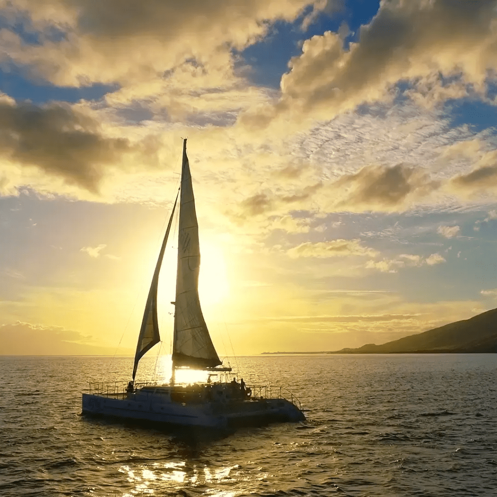 Sailing catamaran on the ocean at sunset with clouds and hills in the background.