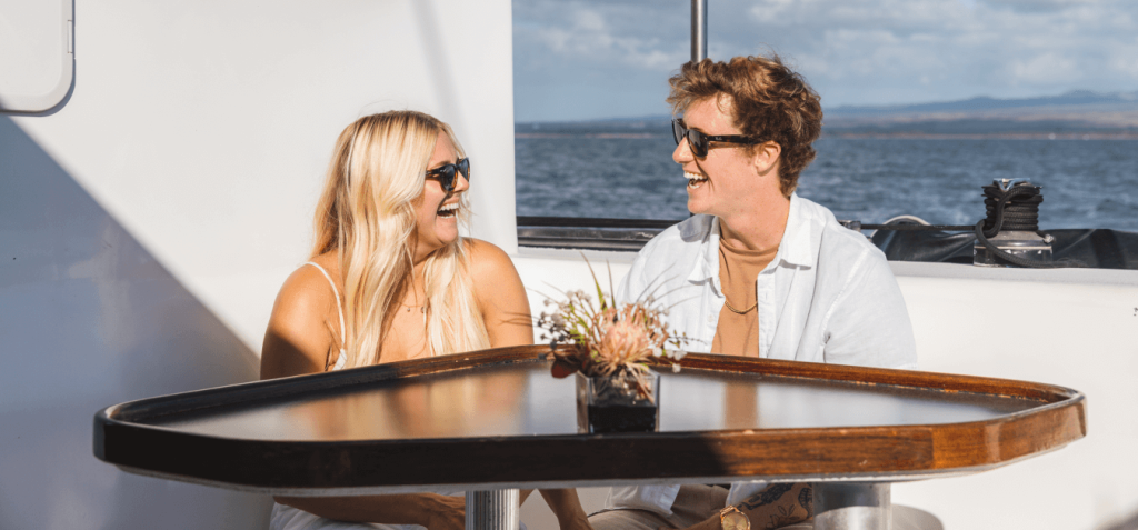 Two people smiling at each other on a boat deck, seated at a table with a sea view.